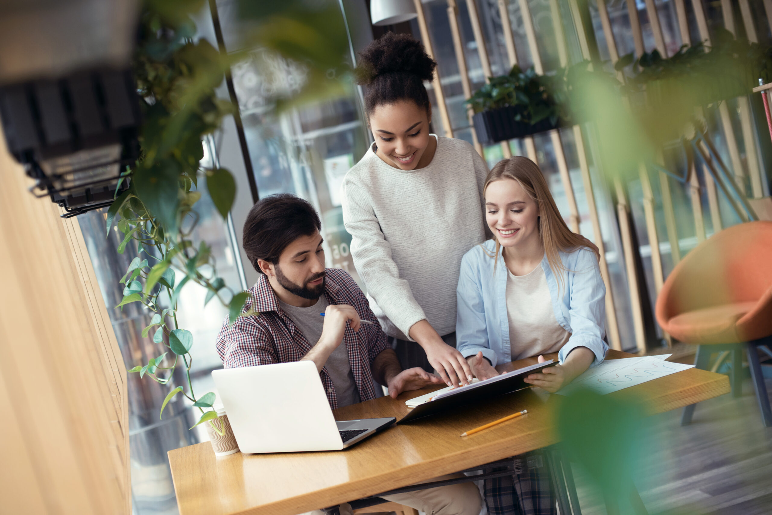 Young people man and women coworking together at creative stylish office sitting at the table african woman explaining data performance pointing at documents on paper holder smiling inspired.